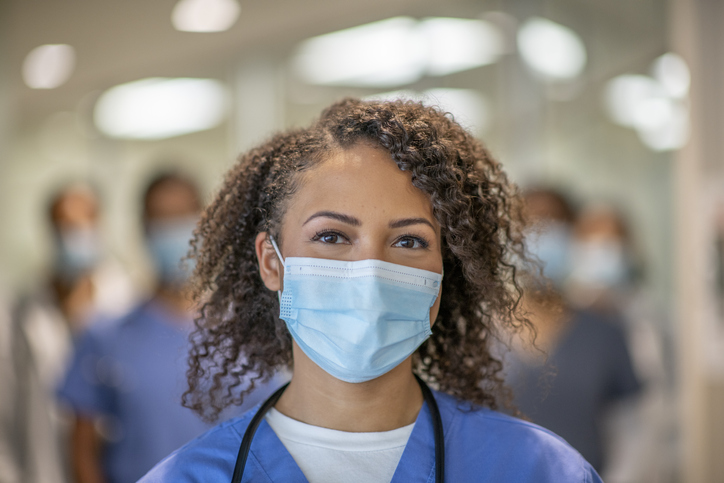 A female doctor is wearing blue medical scrubs and a face mask stands in front of her colleagues at the hospital. She is smiling behind her mask.